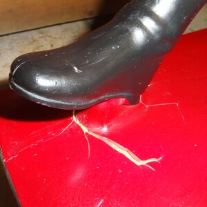 May include: A close-up of a red toy with a black boot stepping on it. The toy has a crack in the red paint and a piece of yellow tape.