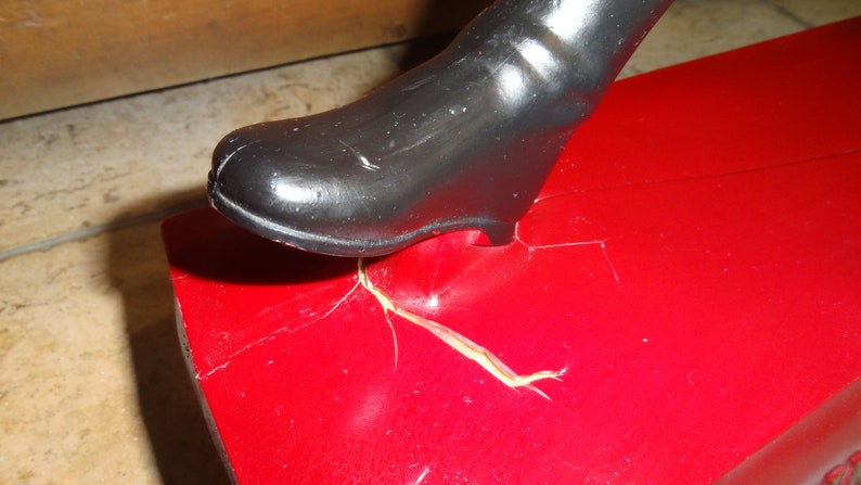 May include: A close-up of a red toy with a black boot stepping on it. The toy has a crack in the red paint and a piece of yellow tape.