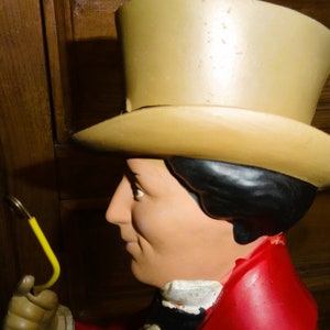 May include: A close-up of a large, colorful, whimsical statue of a person wearing a tall, tan top hat and a red jacket. The statue is holding a yellow object in its hand.