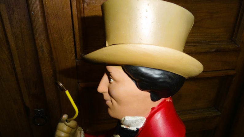 May include: A close-up of a large, colorful, whimsical statue of a person wearing a tall, tan top hat and a red jacket. The statue is holding a yellow object in its hand.
