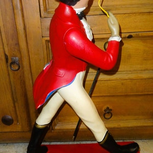 May include: A red and white figurine of a man wearing a brown top hat and a red jacket. He is holding a cane and a monocle in his right hand. The figurine is standing on a red base.