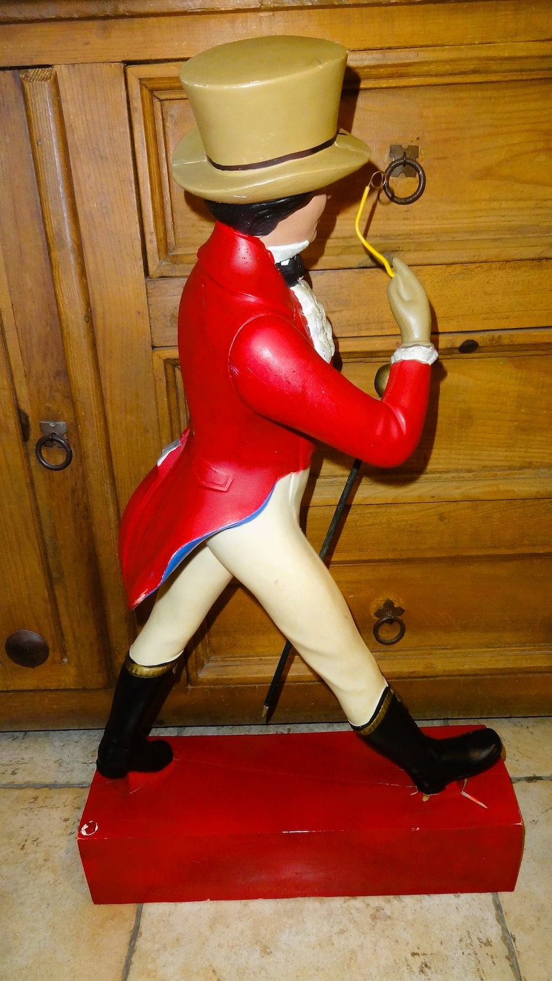 May include: A red and white figurine of a man wearing a brown top hat and a red jacket. He is holding a cane and a monocle in his right hand. The figurine is standing on a red base.