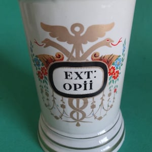 Może przedstawiać: Biały ceramiczny słoik z pozłacanym rantem i złotym i czerwonym wzorem kwiatowym. Słoik ma czarną etykietę z napisem "EXT: OPII" białymi literami.