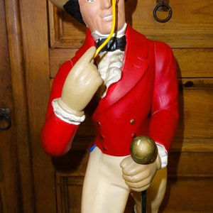 May include: A vintage figurine of a man wearing a brown top hat, red jacket, and white pants. He is holding a black cane and has a yellow monocle.
