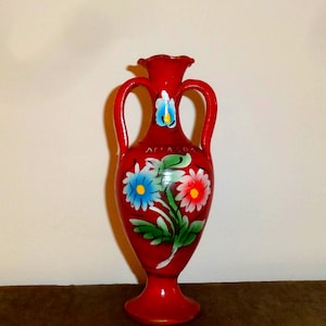 May include: A bright red ceramic amphora vase featuring two handles and a ruffled rim. The body is hand-painted with a floral motif consisting of a blue flower and a red flower with green leaves. The text "ΑΓΙΑ ΣΟΦΙΑ" is visible on the upper body.
