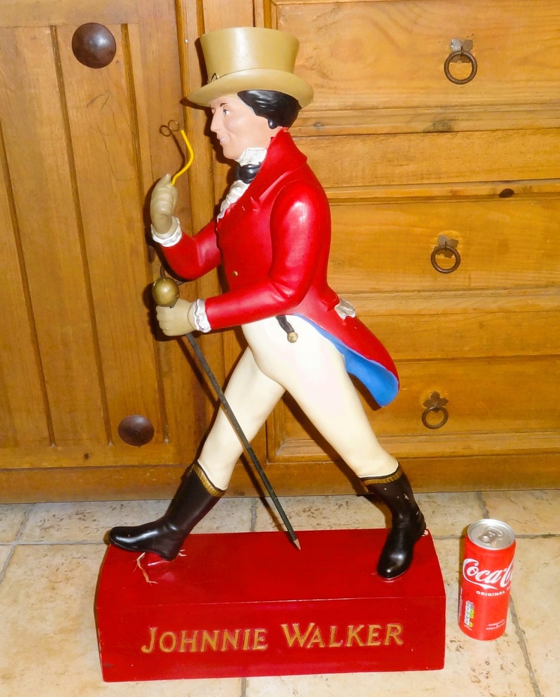 May include: A red, white, and blue figurine of a man in a top hat and red coat holding a walking stick. The figurine is standing on a red base with the words "JOHNNIE WALKER" in gold lettering.
