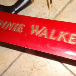 May include: A red metal sign with the words "JOHNNIE WALKER" in gold lettering.