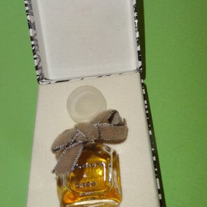 May include: A small, square perfume bottle filled with amber liquid, adorned with a brown bow, sits inside a white box with a black and white patterned border. The bottle's cap is placed next to it.
