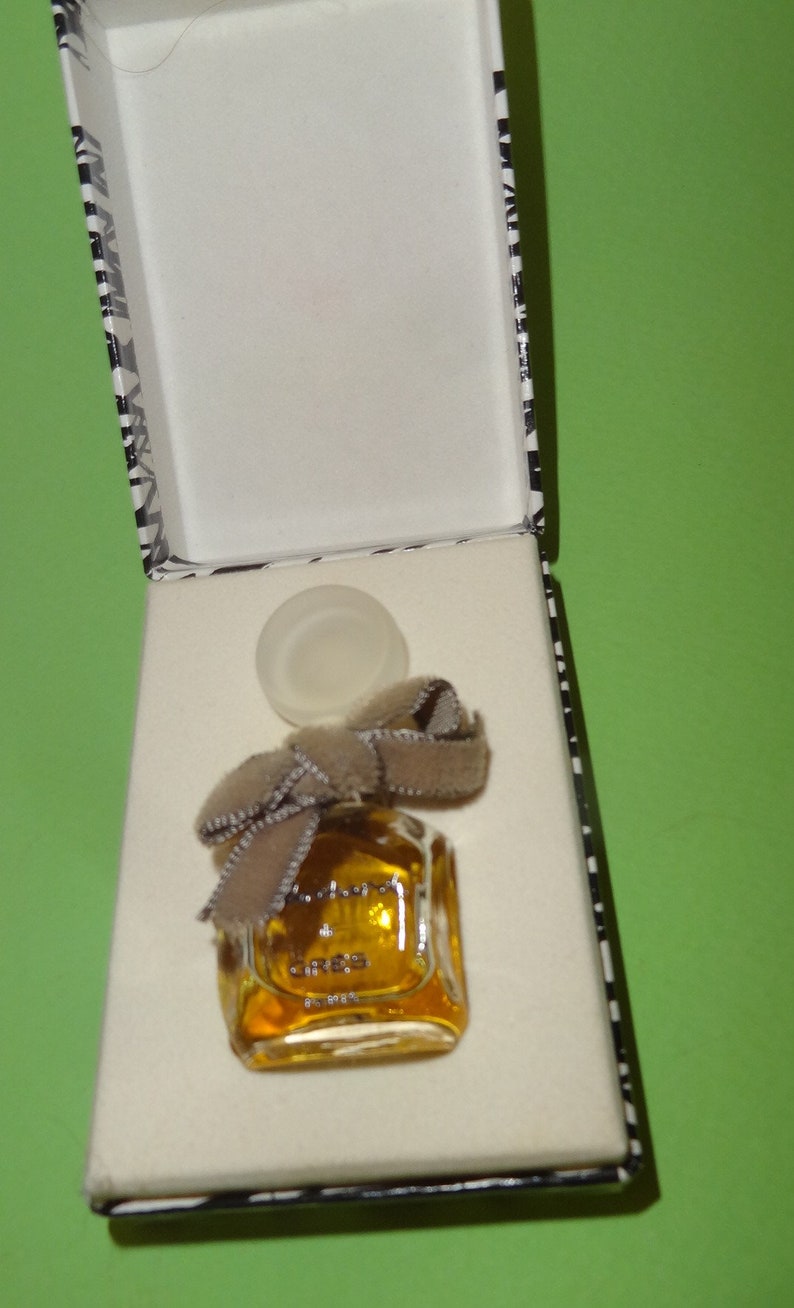 May include: A small, square perfume bottle filled with amber liquid, adorned with a brown bow, sits inside a white box with a black and white patterned border. The bottle's cap is placed next to it.