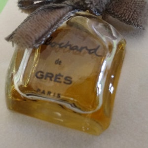 May include: A vintage perfume bottle with a square shape and a light amber hue. The bottle is labeled "Bouchard de GR&Egrave;S PARIS" in black script. A brown ribbon and bow adorn the top of the bottle, adding a touch of elegance.