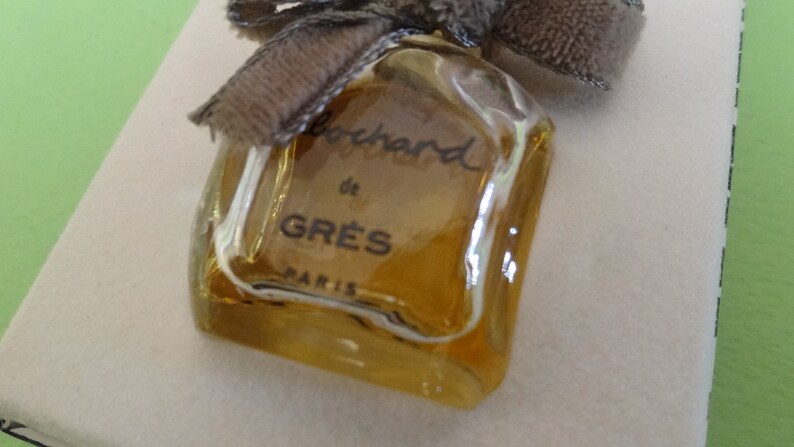 May include: A vintage perfume bottle with a square shape and a light amber hue. The bottle is labeled "Bouchard de GR&Egrave;S PARIS" in black script. A brown ribbon and bow adorn the top of the bottle, adding a touch of elegance.