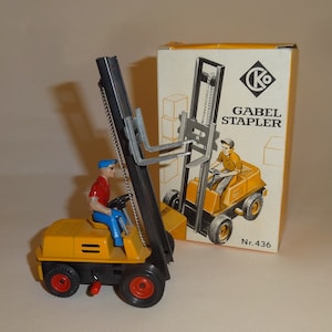 Op de afbeelding: Een gele speelgoed-heftruck met een zwarte mast en een ketting. De heftruck heeft een klein figuurtje van een arbeider in een rood shirt en een blauwe pet die hem bedient. De doos heeft de tekst "Gabel Stapler Nr. 436".