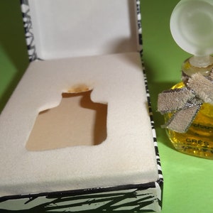 May include: A small, square perfume bottle filled with yellow liquid, adorned with a frosted glass stopper and a beige ribbon. The bottle sits next to an open box with a black and white zebra print design.