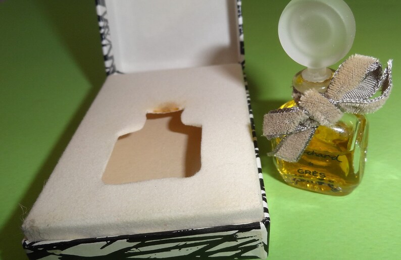May include: A small, square perfume bottle filled with yellow liquid, adorned with a frosted glass stopper and a beige ribbon. The bottle sits next to an open box with a black and white zebra print design.