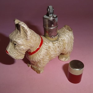 May include: White ceramic figurine of a Scottish terrier with a red collar, holding a silver and chrome lighter. The lighter is separate and has a red and white striped base.