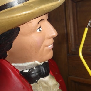 May include: A figurine of a man wearing a brown top hat, a red jacket, and a white shirt with a black bow tie. He is holding a yellow cane with a metal loop at the top.
