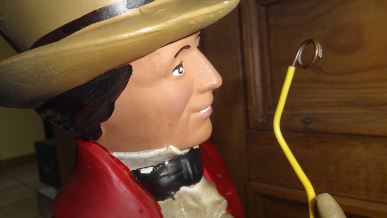 May include: A figurine of a man wearing a brown top hat, a red jacket, and a white shirt with a black bow tie. He is holding a yellow cane with a metal loop at the top.