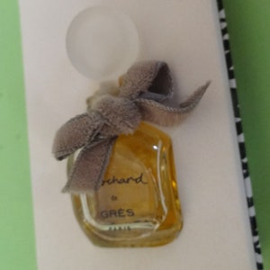May include: A miniature perfume bottle with a clear, rounded stopper and a gray velvet bow. The bottle is filled with a golden liquid and is labeled "orchard de GRES PARIS". The bottle is presented on a white surface.