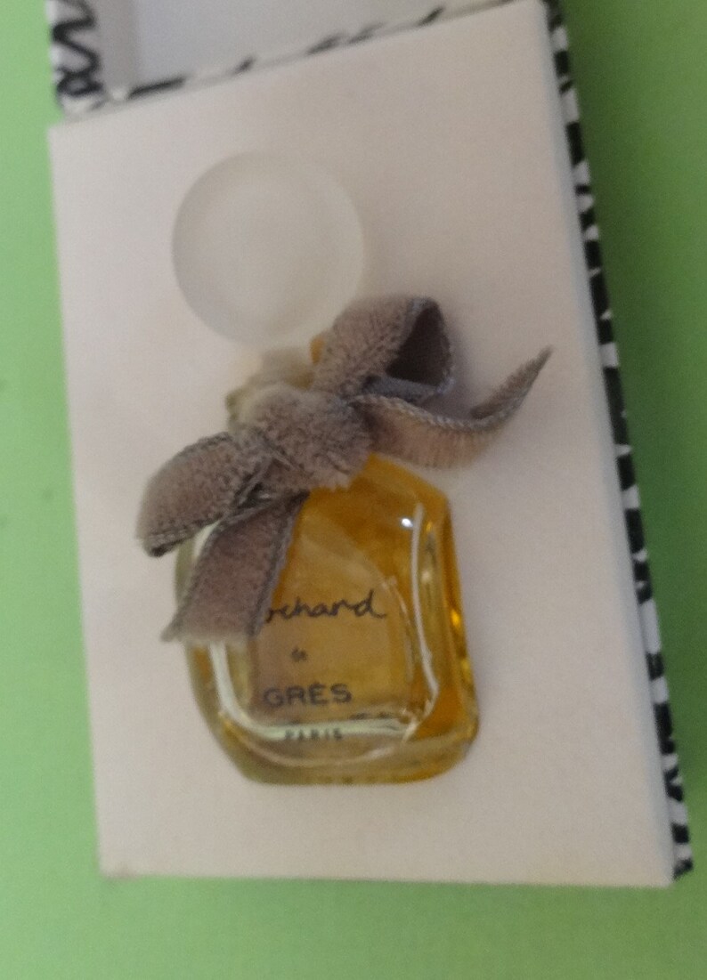 May include: A miniature perfume bottle with a clear, rounded stopper and a gray velvet bow. The bottle is filled with a golden liquid and is labeled "orchard de GRES PARIS". The bottle is presented on a white surface.