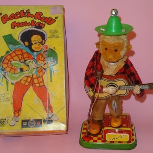 May include: A vintage tin toy monkey wearing a green hat and plaid shirt plays a guitar. The monkey rocks back and forth as it plays. The toy is battery operated and has a lighted microphone. The box reads "Rock 'n' Roll Monkey".