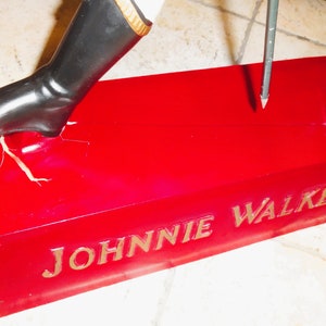 May include: A red base with the words "JOHNNIE WALKER" in gold lettering. A black boot is stepping on the base, and a metal rod is sticking into the base.