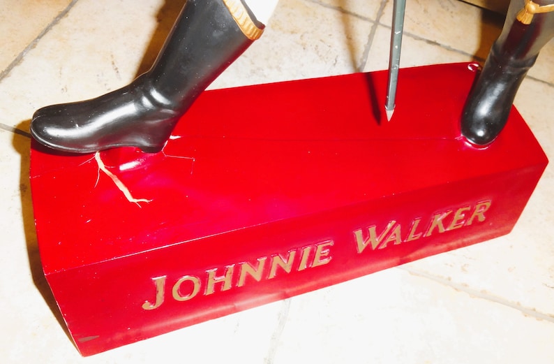 May include: A red base with the words "JOHNNIE WALKER" in gold lettering. A black boot is stepping on the base, and a metal rod is sticking into the base.