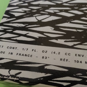 May include: A rectangular box with a black and white abstract design. The text on the box reads "NET CONT. 1/7 FL. OZ (4.2 CC ENV.) MADE IN FRANCE - 83&deg; - R&Eacute;F. 104 N". The box is on a green surface.