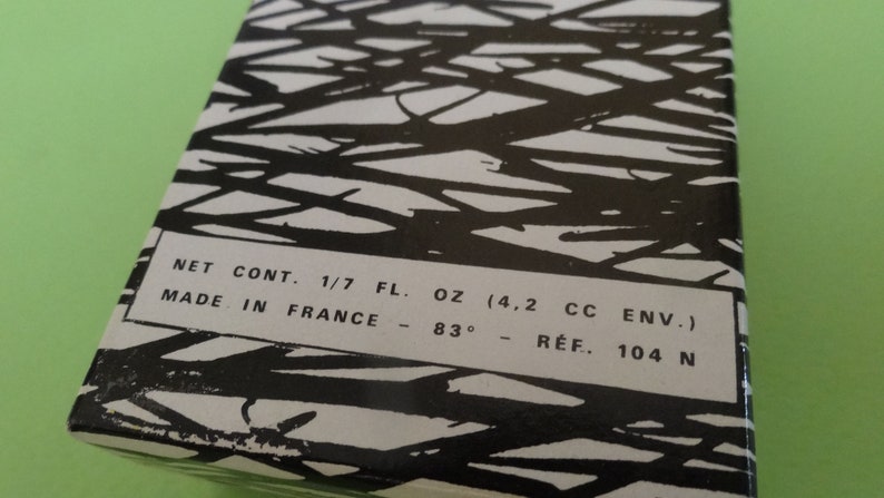 May include: A rectangular box with a black and white abstract design. The text on the box reads "NET CONT. 1/7 FL. OZ (4.2 CC ENV.) MADE IN FRANCE - 83&deg; - R&Eacute;F. 104 N". The box is on a green surface.
