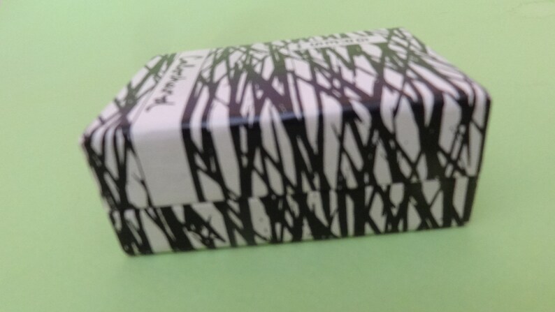 May include: A rectangular box with a white background and a black tree branch pattern. The box has a hinged lid and a white vertical stripe on the front. The box is on a light green surface.