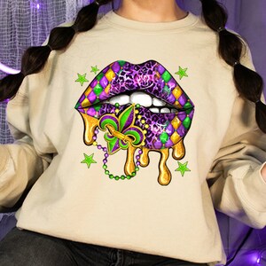 Op de afbeelding: Een beige sweatshirt met een Mardi Gras-thema lippenontwerp in paars, groen en goud. De lippen druipen met gouden verf en hebben een fleur-de-lis in het midden.