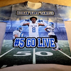 May include: A blue and white sublimated t-shirt with a football player in the center. The shirt has the text "Keiran "Hawk" Daniels" and "#5 Go Live" on it. The background is a football stadium.