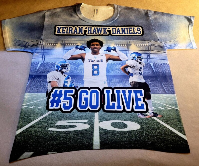 May include: A blue and white sublimated t-shirt with a football player in the center. The shirt has the text "Keiran "Hawk" Daniels" and "#5 Go Live" on it. The background is a football stadium.