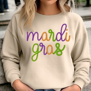 Op de afbeelding: Een beige sweatshirt met de woorden "Mardi Gras" geborduurd in paars, groen en oranje garen.
