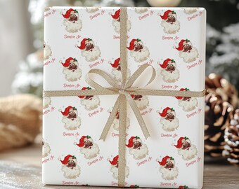 May include: White gift wrap with a repeating pattern of a cartoon Santa Claus with a black face and the name "Denton Jr." printed in red. The gift wrap is tied with a gold ribbon.