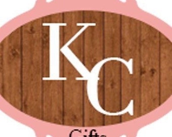 May include: A pink oval frame with a woodgrain background. The letters "KC" are in white against the woodgrain. The text "Gifts" is below the letters in white.