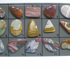 May include: A display of 16 polished agate stones, each with unique patterns and colors, including red, brown, white, and green. The stones are arranged in a grid on a gray background.