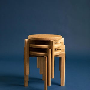 Könnte beinhalten: Ein Stapel aus vier Holzstühlen mit natürlicher Oberfläche. Die Hocker haben runde Sitze und quadratische Beine, mit einem minimalistischen Design. Der Hintergrund ist einfarbig blau.