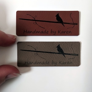 May include: Two brown leather labels featuring a black silhouette of a cardinal perched on a branch with a needle. The text "Handmade by Karen" is printed on each label.