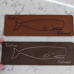 May include: Two brown leather coasters with a white whale outline and the text "Call me Ishmael" on each. The top coaster is brown and the bottom coaster is a darker brown.