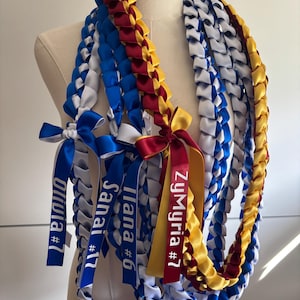 May include: A collection of braided satin ribbon leis in blue, white, gold, and maroon. Each lei features a ribbon bow with printed text including the names Olivia #7, Sanai #17, Ilana #6, and ZyMyria #7, displayed on a mannequin torso.