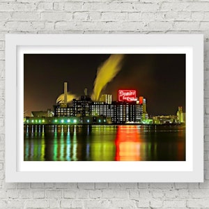 May include: A nighttime view of a large industrial building with a red neon sign that reads "Domino Sugar" and smoke coming from the building's chimneys. The building is reflected in the water in front of it.