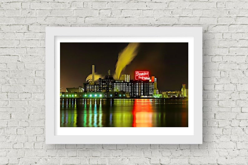 May include: A nighttime view of a large industrial building with a red neon sign that reads "Domino Sugar" and smoke coming from the building's chimneys. The building is reflected in the water in front of it.