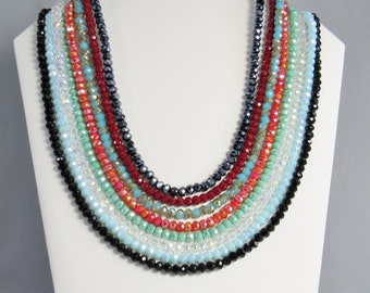 May include: A multi-strand necklace made of faceted glass beads in various colors, including black, red, turquoise, and clear. The necklace is layered and creates a cascading effect.