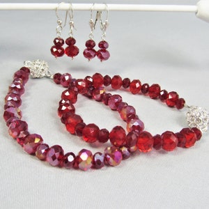 May include: A set of red crystal jewelry featuring a necklace, bracelet, and earrings. The necklace and bracelet have a silver clasp with clear rhinestones. The earrings are silver with red crystal drops.