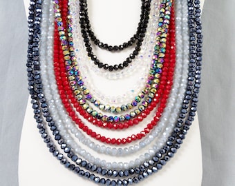 May include: A multi-strand necklace featuring faceted crystal beads in a layered design. The strands include colors of black, clear, iridescent, red, light gray, and dark blue, arranged in a cascading pattern on a white display form.