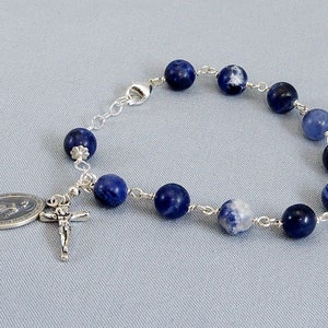 May include: A silver bracelet with blue and white beads and a silver charm with a religious image. The bracelet is made of silver chain and has a clasp.