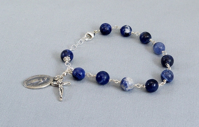 May include: A silver bracelet with blue and white beads and a silver charm with a religious image. The bracelet is made of silver chain and has a clasp.