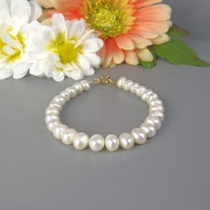 May include: A white pearl bracelet with a gold clasp. The bracelet is composed of round, lustrous pearls. White and orange flowers are in the background. The bracelet is a classic piece of jewelry.
