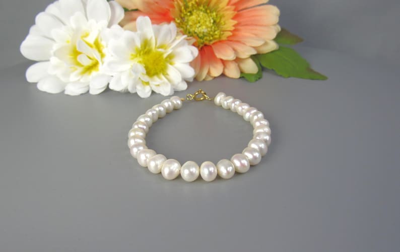 May include: A white pearl bracelet with a gold clasp. The bracelet is composed of round, lustrous pearls. White and orange flowers are in the background. The bracelet is a classic piece of jewelry.