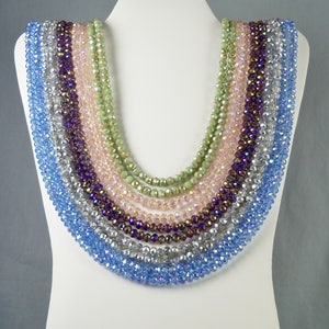 May include: A set of seven crystal necklaces in various shades of blue, green, and purple. The necklaces are layered on a white mannequin bust.